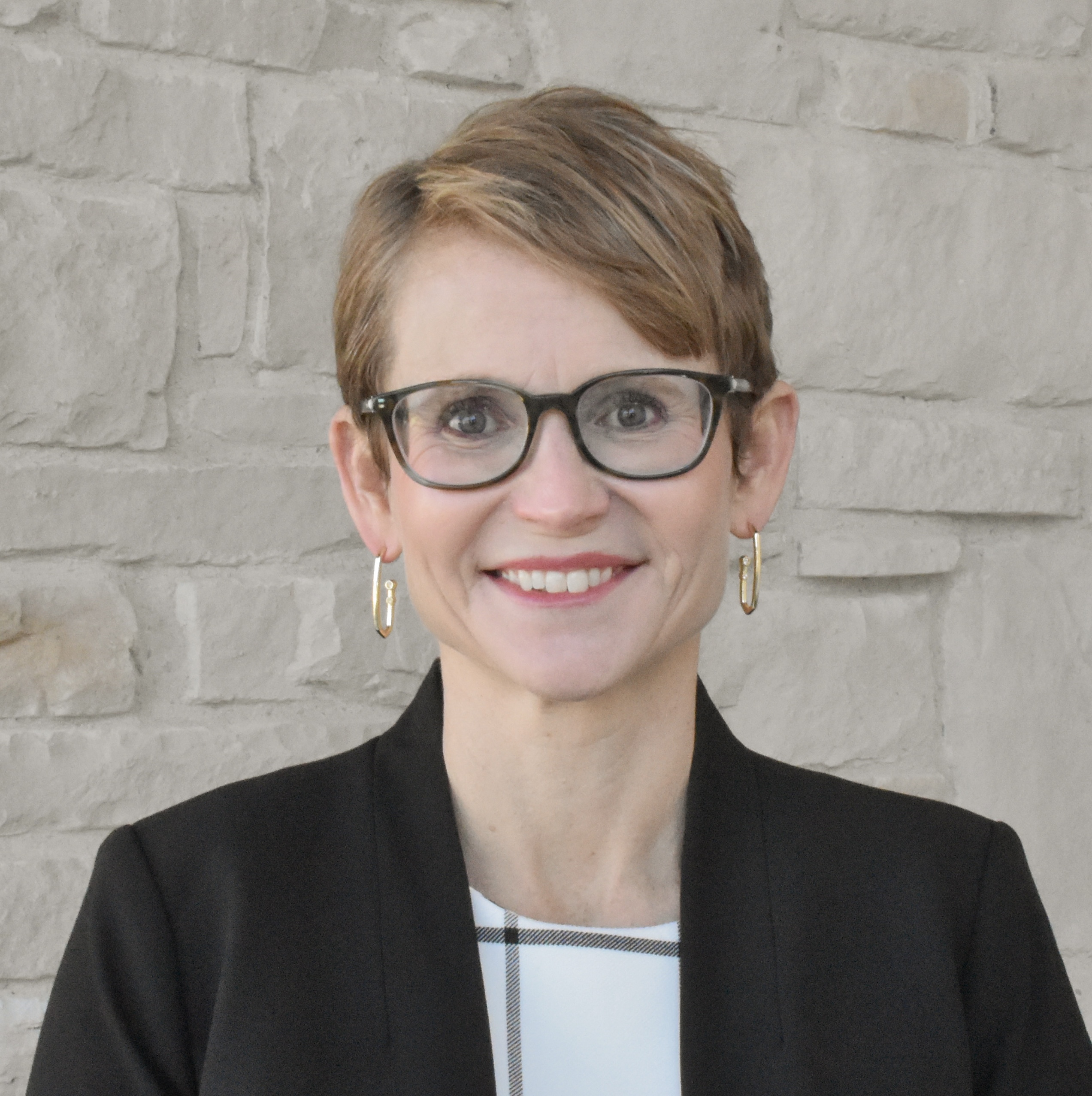 a headshot of a woman with short light brown hair in black glasses and geometric earrings who is standing in front of a light rock wall in a black blazer over a white and black shirt.